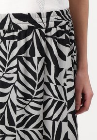 Jupe à motif de feuilles noir et blanc avec une ceinture élastique portée avec un haut blanc, montrant la main droite d'une personne sur le côté.
