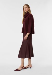 Frau in einem bordeauxfarbenen Rollkragenpullover, passendem Midikleid und flachen Slingback-Schuhen, die seitlich vor einem weißen Hintergrund steht.