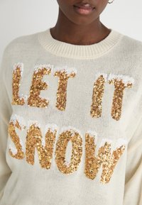 Krämfärgad stickad tröja med stora guld- och vitglittriga bokstäver som står "LET IT SNOW." Rund halsringning och texturerad yta.