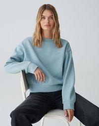 Felpa azzurra con scollo rotondo e polsini a coste, con un piccolo logo a forma di cuore. Jeans neri, seduto su una sedia.