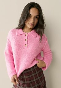 Rožinė pūkuota megztinė su apykakle, trimis mediniais sagomis ir ilgomis rankovėmis. Dėvima virš rudos languotos sijono su kelnių ilgio apkarpymu.