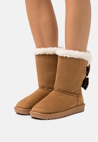 Bottines en daim marron avec un revers en fourrure blanche, présentant un détail de nœud lacé sur le côté et une semelle en caoutchouc texturé pour une meilleure adhérence.