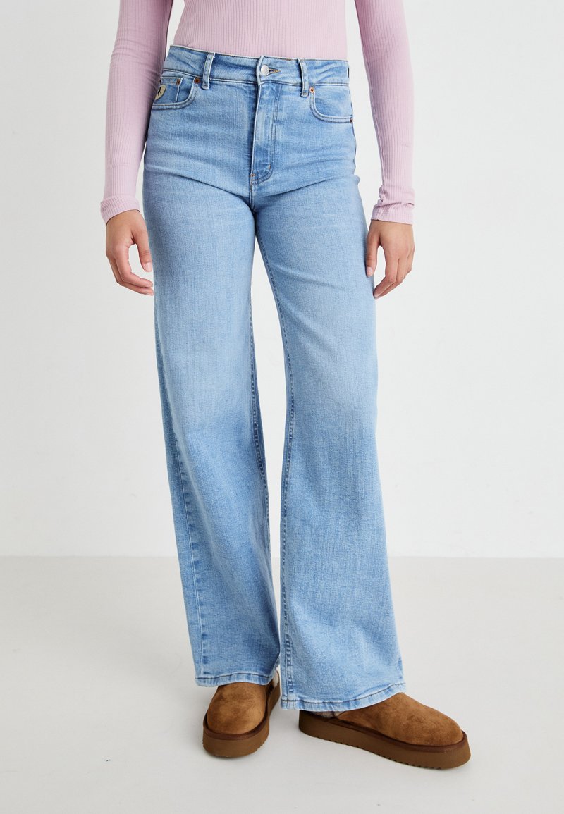 Person som har på seg lyseblå vide jeans, brune semskede slip-on sko, og en langermet ribbet rosa topp mens de står på en ren hvit bakgrunn.