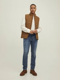 Braune gesteppte Weste über einem weißen Langarmshirt, kombiniert mit blauen Jeans und braunen Schnürschuhen. Model steht vor einem neutralen Hintergrund.