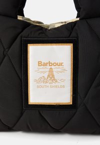Schwarze gesteppte Tasche mit einem beige- und schwarzkarierten Innenfutter sowie einem weißen Patch, auf dem ein Leuchtturm und der Text "Barbour South Shields" zu sehen sind.