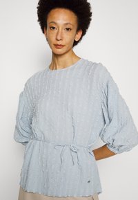 Blusa azul clara com riscas verticais texturizadas, mangas bufantes e cintura com cordão. Terminada com um pequeno detalhe de logótipo próximo da bainha.