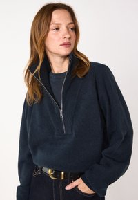 Maglione zip-up blu navy con colletto alto, realizzato in materiale morbido. Caratterizzato da un collo a coste e maniche ampie e rilassate. Jeans in denim con cintura.