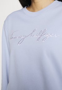 Sweat-shirt bleu clair avec le logo "Tommy Hilfiger" brodé en blanc. Texture douce et encolure côtelée. Vue en gros plan.