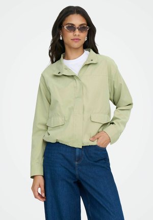 Žena v svetlo zelenej mikine na zips, bielom tričku, modrých džínsoch, strieborných náušniciach a slnečných okuliaroch s mačacími očami, stojaca s jednou rukou v vrecku.