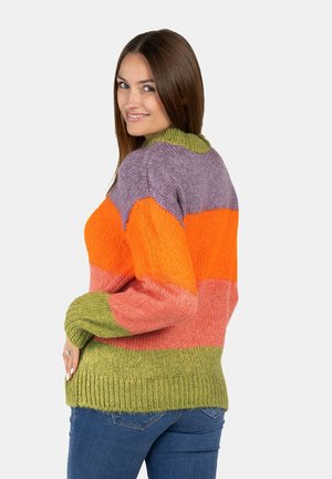 Ung kvinde smiler, iført en flerfarvet strikket sweater med grønne, pink, orange og lilla striber, kombineret med blå jeans.