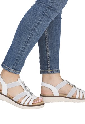 Fødder med hvide sandaler med stropper og sølvspænder kombineret med blå skinny jeans mod en hvid baggrund.