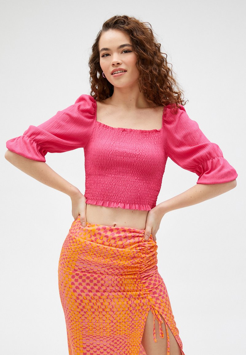 Lyserød cropped bluse med smocked tekstur og puffærmer, parret med en livlig mønstret nederdel med cirkler i lyserøde og orange nuancer.