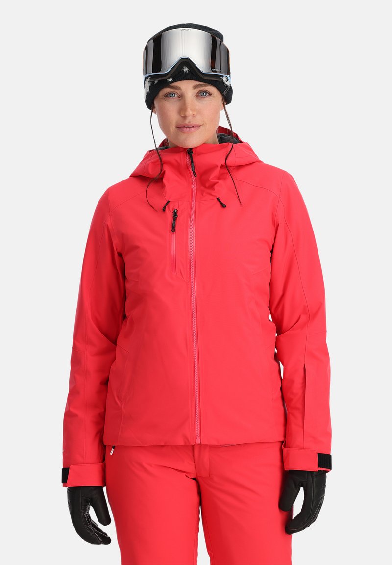 Helle pinke Skijacke mit Reißverschluss, verstellbarer Kapuze und Reißverschlusstaschen. Schwarze Handschuhe und spiegelnde Skibrille runden den Look ab.