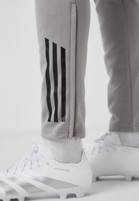 Grijze trainingsbroek met een zwarte drie-strepen ontwerp aan de zijkant, gecombineerd met witte voetbalschoenen met zilveren accenten en een gestructureerde oppervlakte.
