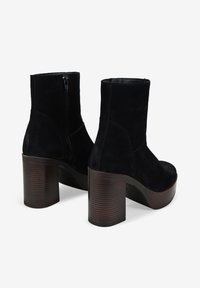 Botines de ante negro con cremalleras laterales y gruesos tacones de bloque de madera apilados, mostrados desde la parte trasera y en vista en ángulo.