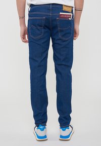 Jeans in denim blu scuro con cuciture arancioni, due tasche posteriori e un'etichetta di marca. Indossati con sneakers casual grigie e blu.