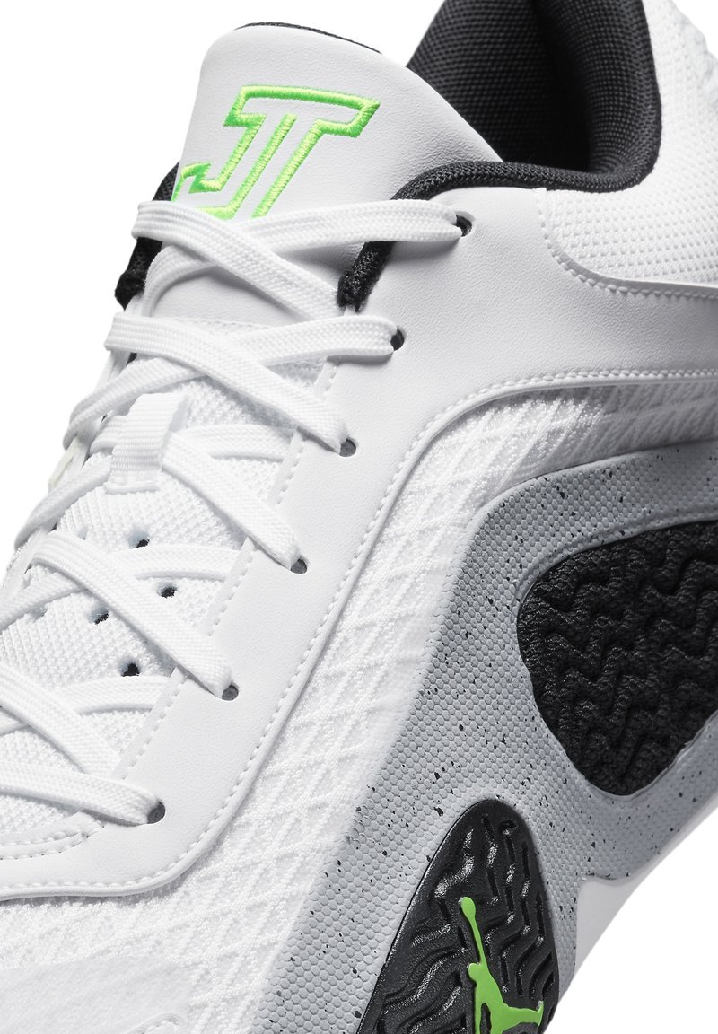 Zapatilla deportiva blanca con acentos verdes y negros, con un lateral texturizado en gris, un logo en la lengua, y cordones blancos con detalles de malla.