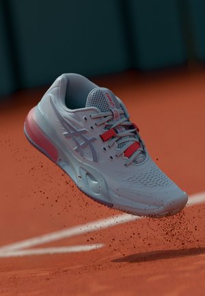 Zapatilla de tenis azul claro con parte superior texturizada, detalles en rojo y una suela semitransparente, mostrada en el aire sobre una superficie de arcilla.