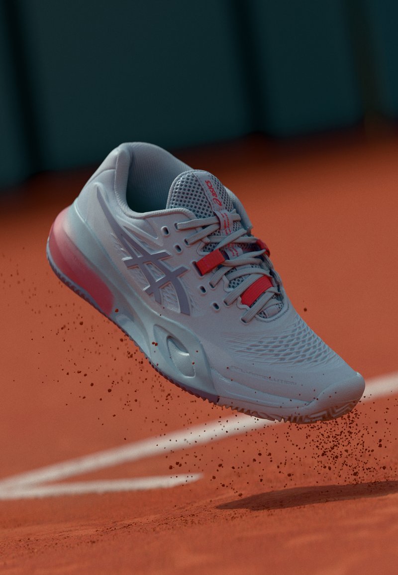 Chaussure de tennis bleu clair avec une tige texturée, des accents rouges et une semelle semi-transparente, montrée en l'air au-dessus d'une surface en terre battue.