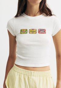 Valkoinen lyhythihainen t-paita, jossa on kolme värikästä sardiinipurkin grafiikkaa. Pehmeä puuvillamateriaali, jossa on slim fit -malli ja lyhyet hihat.