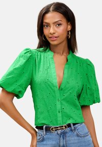 Donna che indossa una camicetta verde brillante con maniche corte a sbuffo e bottoni, caratterizzata da un motivo floreale a occhielli, abbinata a jeans blu e orecchini a cerchio dorati.