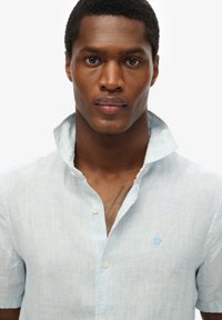 Lichtblauw, kort mouwen overhemd met kraag en een knoopsluiting, voorzien van dunne witte strepen en een klein logo op de borst.