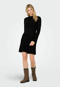 Frau mit langen blonden Haaren, die ein schwarzes ribbeliges Langarmkleid und braune Stiefel bis zur Mitte der Waden mit grauen Socken trägt, stehend vor einem weißen Hintergrund.