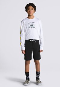 Camicia bianca a maniche lunghe con logo sul petto e design di faccine sorridenti gialle. Pantaloni corti neri con dettagli a scacchi sui lati. Scarpe slip-on a scacchi in bianco e nero.