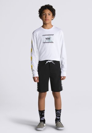 Witte lang-voor mouw shirt met borstlogo en gele glimlach ontwerpen. Zwarte shorts met een zwart-wit geblokt accenten aan de zijkant. Zwart-witte geblokte slip-ons.
