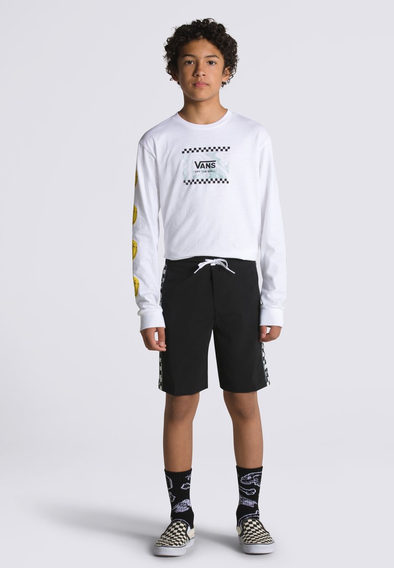 Camicia bianca a maniche lunghe con logo sul petto e design di faccine sorridenti gialle. Pantaloni corti neri con dettagli a scacchi sui lati. Scarpe slip-on a scacchi in bianco e nero.