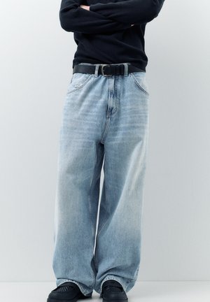 Osoba ubrana w jasnoniebieskie jeansy z szerokimi nogawkami, czarny pasek, czarną bluzkę z długim rękawem oraz czarne buty, stojąca z założonymi rękami.