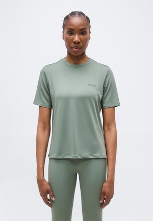 Γυναίκα με πλεξούδες, φορώντας ένα ανοιχτό πράσινο κοντομάνικο πουκάμισο και ταιριαστά κολάν, στέκεται μπροστά σε απλό ανοιχτό φόντο.