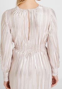 Robe à manches longues en rose pâle, dotée de plis verticaux, d'une taille smockée et d'un dos en trou de serrure avec un bouton de fermeture. Texture satin lisse.