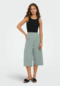 Czarna prążkowana krótka bluzka, wysoka talia jasnozielonych spodni w szerokim kroju z plisami oraz czarne klapki z kwiatowymi akcentami.