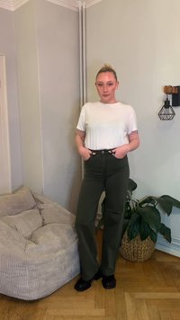 Bijela majica kratkih rukava uparena s visokim strukom maslinasto zelenih hlača s prednjim džepovima, nošena s crnim cipelama. Pozadina od mekog, teksturiranog kauča i biljke.