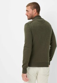 Gr�üner Strickpullover mit hohem Kragen und gerippten Manschetten, der eine strukturierte Oberfläche und eine taillierte Passform aufweist. Getragen mit hellen Hosen.