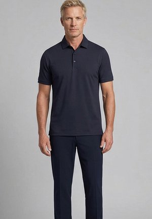 Man met kort blond haar, gekleed in een zwart poloshirt met korte mouwen en een zwarte broek, staand tegen een eenvoudige grijze achtergrond.