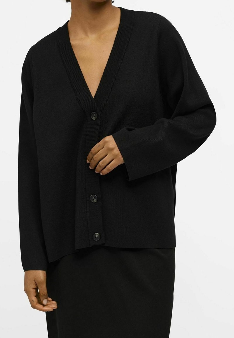 Personne portant un cardigan noir à col en V boutonné avec des manches longues et une jupe noire assortie sur un fond blanc.