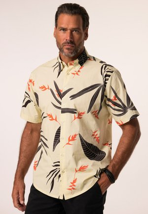 Man met kort donker haar en baard, die een beige shirt met korte mouwen draagt met zwarte en oranje tropische bladpatronen en een zwarte broek.