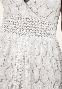 Weißes Spitzenkleid mit floralen Mustern, strukturiertem Bund und feinen Details, einem schichtweise aufgebauten Rock und aufwendigen Spitzenakzenten.
