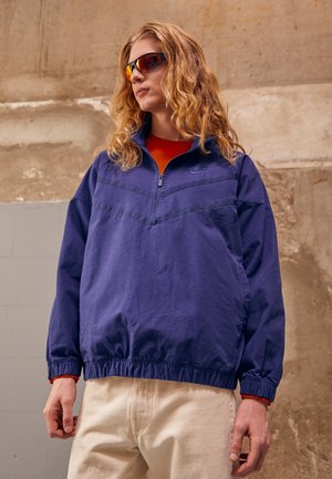 Marineblå pulloverjakke med halv lynlås, elastiske manchetter og kant. Orange accent på den indvendige krave og logo på brystet. Båret med beige bukser.