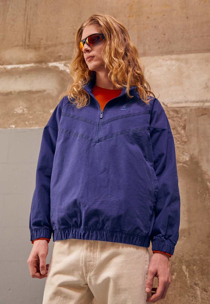 Tumši zila puloverjaka ar pusvelkamu rāvējslēdzēju, elastīgām piedurknēm un apakšmalu. Oranža akcenta detaļa iekšējā apkaklē un logo uz krūts. Valka kopā ar bēšiem biksēm.