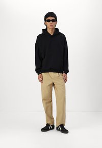Schwarzer Kapuzenpullover mit Kängurutasche, beigefarbene Hose und schwarze Sneakers mit weißen Streifen, getragen von einer Person mit Sonnenbrille.