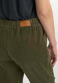 Close-up van een persoon die een olijfgroene corduroy broek met een elastische tailleband draagt, met een bruine leren merketiket aan de achterkant.