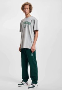 Grå t-shirt med grøn tekst, parret med mørkegrønne bukser med et logo. Sættet er suppleret af sort-hvide sneakers.