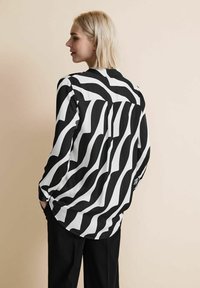 Blusa con motivo ondulato in bianco e nero, maniche lunghe, chiusura a bottoni sul retro e vestibilità ampia, realizzata in un tessuto leggero e morbido.