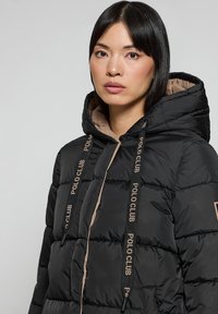Svart puffjacka med beige foder, utrustad med huva, framträdande sydda sektioner och "POLO CLUB"-logotyp på dragskorna.
