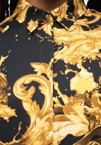Μαύρο πουκάμισο με περίτεχνα χρυσά μοτίβα μπαρόκ σε στυλ λουλουδιών και έλικες, με γιακά με κουμπιά και διακοσμητικό χρυσό κουμπί.