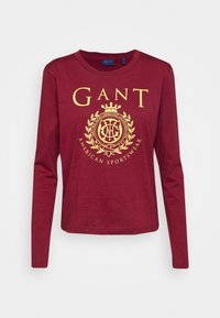 Burgunderfarbendes Langarm-T-Shirt aus Baumwolle, mit einem großen goldenen Logodesign, das auf der Vorderseite zentriert ist. Klassischer Rundhalsausschnitt und gerade Passform.