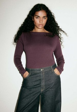 Femme aux longs cheveux bouclés portant un haut violet moulant à manches longues et un jean large taille haute de couleur foncée, les mains dans les poches.